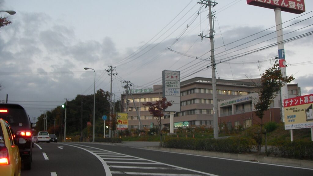 富谷中央病院の建物と前の道路。信号と店舗の看板が並ぶ