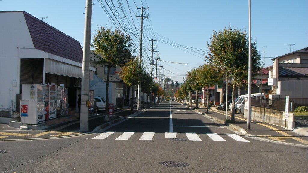 富谷市内の通りに面した個人商店。歩道沿いに自動販売機が並び、まっすぐな道が続く