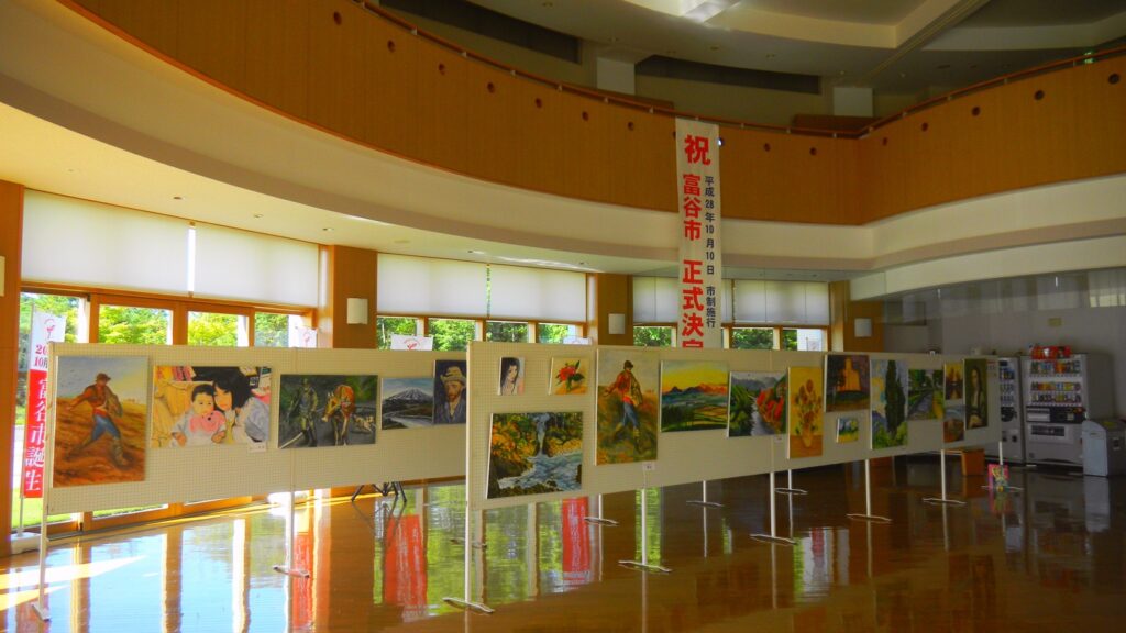 富谷市民交流ホールの市民ギャラリー。曲面の壁に沿って絵画がパネル展示され、「祝　富谷市正式決定」の垂れ幕が下がっている