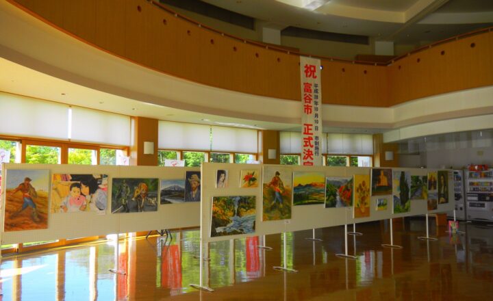 富谷市民交流ホールの市民ギャラリー。曲面の壁に沿って絵画がパネル展示され、「祝　富谷市正式決定」の垂れ幕が下がっている